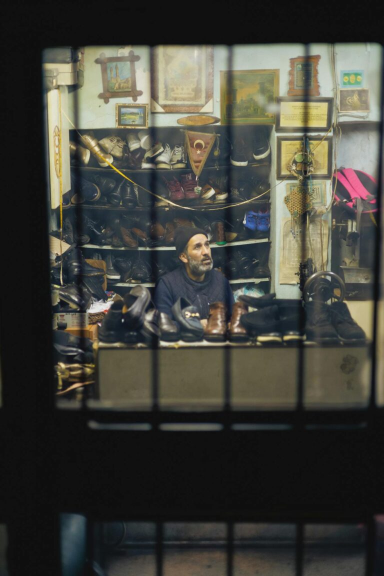 A mature shoemaker in his workshop surrounded by handmade shoes, viewed through a window.