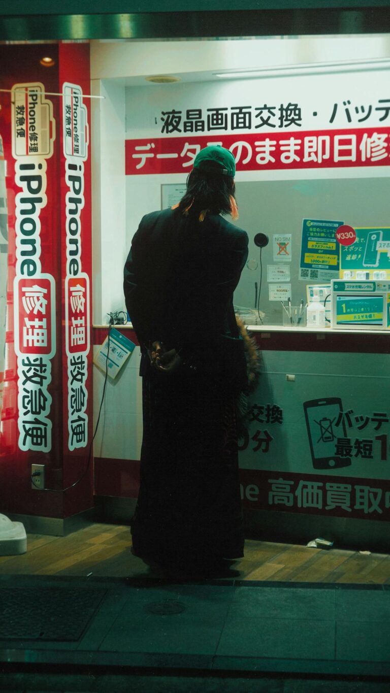 A person in a hat stands at a shop counter with illuminated signage at night.